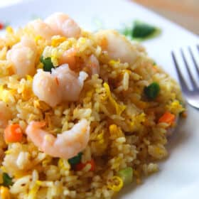 A plate of Healthy Shrimp Fried Rice with mixed vegetables, with green peas and carrot bits, paired with a fork on the side, served on a white plate that is sitting on a wooden table.