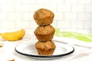 Three stacked Healthy Banana Protein Muffins on a white plate with a black rim. Sitting on a marble countertop with a subway tile background and a banana off to the left.