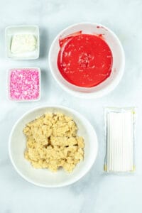 Top view of ingredients on a marble surface, including a bowl of red glaze, a bowl of crumbled dough, white icing, pink sprinkles, and a packet of white sticks. For making Homemade Valentine's Cake Pops.