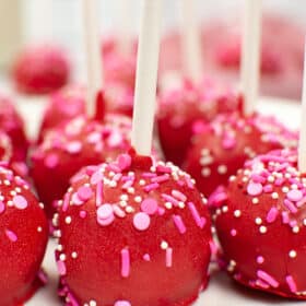 Homemade Valentine's Cake Pops coated with red chocolate and decorated with pink and white sprinkles on lollipop sticks, arranged closely together.
