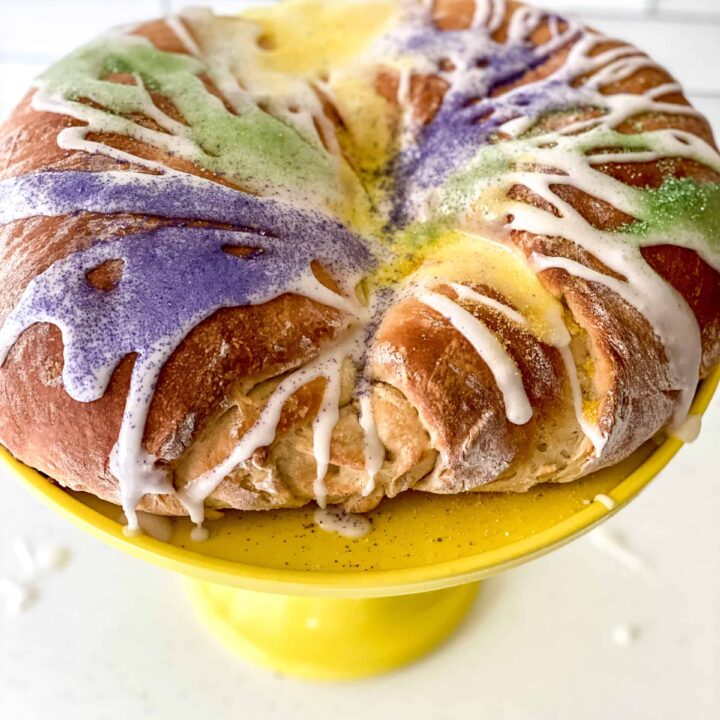 A vibrant king cake with purple, green, and gold sugar toppings and white icing, displayed on a yellow cake stand against a white tiled background.