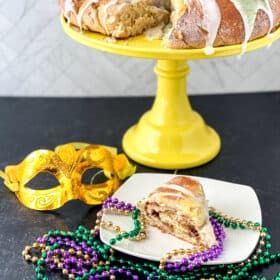 A colorful king cake on a yellow cake stand, with a slice placed on a white plate accompanied by Mardi Gras beads and a golden mask.