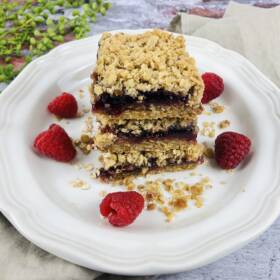 Two Raspberry Oatmeal Bars stacked on a white plate, garnished with fresh raspberries and crumbs, set on a rustic surface with greenery in the background.