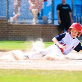A young boy in a white and red baseball uniform, helmet included, sliding into a dusty base during a sunny day at a baseball game.