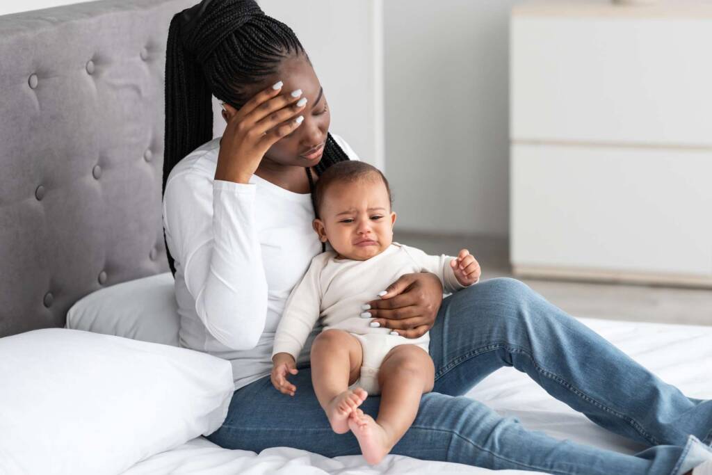 Tired African American young mom sitting with kid on bed