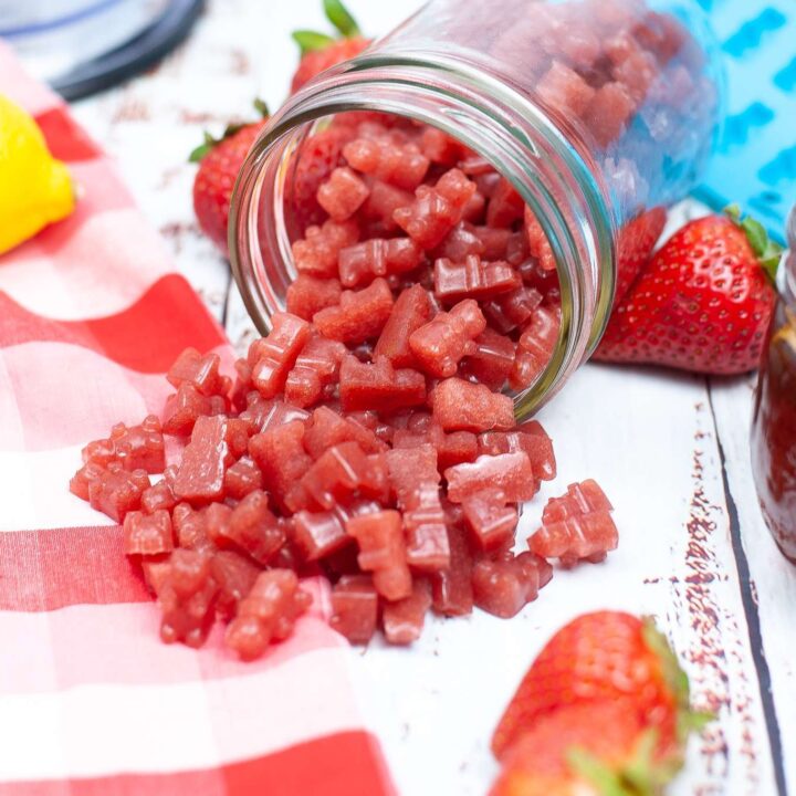 A glass jar tipped over with Healthy Homemade Strawberry Gummies spilling onto a white wooden surface, surrounded by fresh strawberries and a checkered red and white cloth. With a blue silicone gummy bear mold in behind the jar.