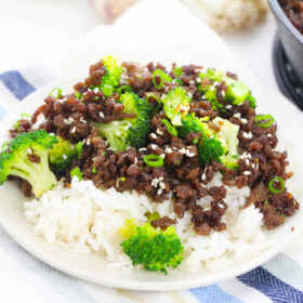 A plate of Easy Korean Beef and Broccoli, garnished with sliced green onions, served besides the skillet it was cooked in/ Situatied on a blue and white striped napkin, with green onions and garlic towards the top of the frame.
