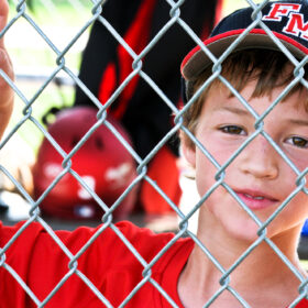 A young boy wearing a red baseball uniform and cap peeks through a chain-link fence, smiling slightly with one hand resting on the fence.