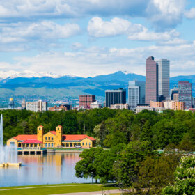 A vibrant view of denver skyline from city park, featuring a fountain in a lake, lush greenery, and snow-capped rocky mountains in the background under a clear blue sky.