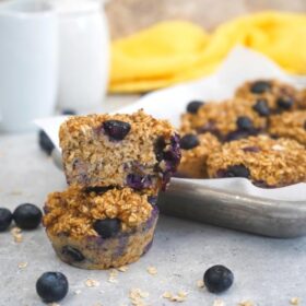 Freshly baked blueberry oat cups on a metal tray with a white cup in the background, surrounded by loose blueberries and a yellow napkin on a textured surface.