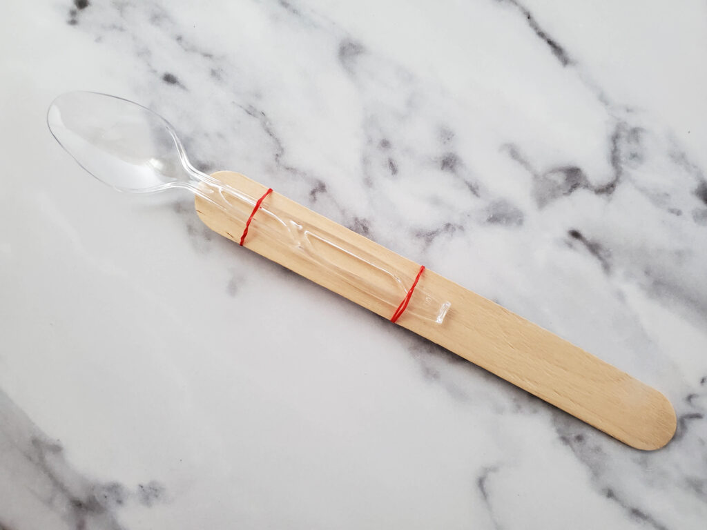 attaching a plastic spoon to a popsicle stick with rubber bands