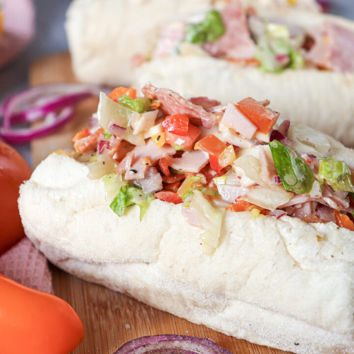 Two fresh Chopped Italian Sandwiches filled with a mix of meats, chopped vegetables, and dressing on a wooden board, surrounded by whole tomatoes and sliced red onions.