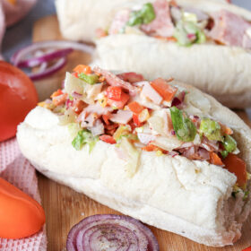 Two fresh Chopped Italian Sandwiches filled with a mix of meats, chopped vegetables, and dressing on a wooden board, surrounded by whole tomatoes and sliced red onions.