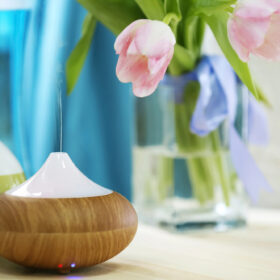 An essential oil diffuser with a wooden base and white top emitting vapor, sitting on a table with blurred pink flowers in the background.