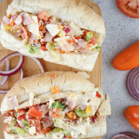 Two Chopped Italian Sandwiches filled with fresh diced vegetables, meat, and cheese on a wooden cutting board surrounded by sliced tomatoes, onions, and peppers.